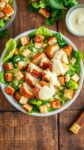 a colorful bowl of caesar salad with crispy chicken, lettuce, croutons, and parmesan on a wooden table.